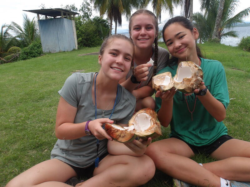 Three young women are sitting on the grass, each holding a halved coconut. They are smiling at the camera. The coconuts are cracked open, revealing the white flesh inside. In the background, there's a small building and some palm trees, suggesting a tropical location.
