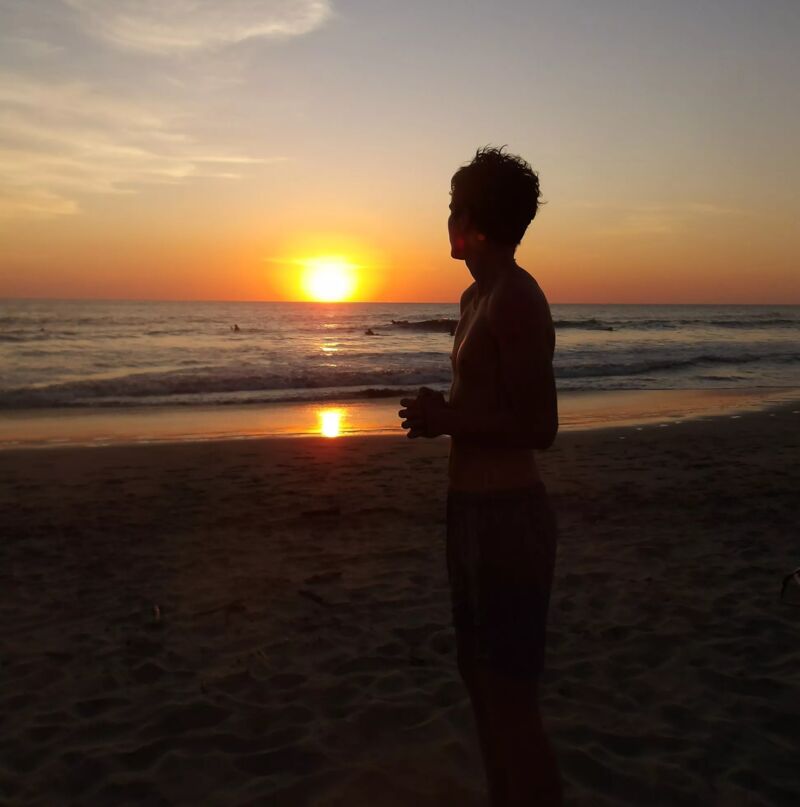 A person stands on a sandy beach, gazing out at a vibrant sunset over the ocean. The sun reflects on the water, creating a shimmering path. The sky is painted with warm hues of orange and yellow. The individual is silhouetted against the bright light, adding a sense of contemplation and serenity to the scene. The waves gently roll onto the shore, completing the tranquil coastal setting.
