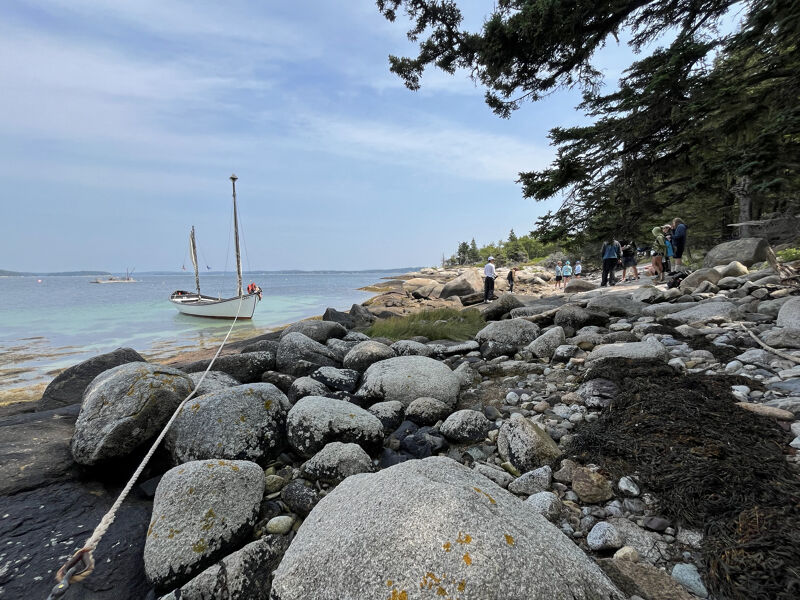 The image shows a rocky beach with a sailboat in the background. The sky is cloudy and the water is calm. There are people walking on the beach in the distance. The foreground is filled with large rocks and seaweed. Trees line the edge of the beach on the right side of the image.