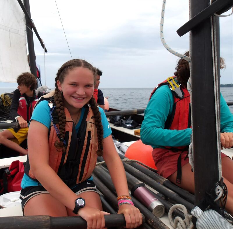 A group of people are on a sailboat. A young girl with braided hair is in the foreground, wearing a life jacket and holding an oar. Other people are visible in the background, also wearing life jackets. The boat is on the water, and the sky is overcast.