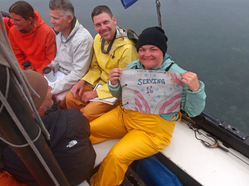 A group of people are on a boat, possibly fishing or touring. The person in the foreground is holding up a sign that says "Serving 16". They are wearing yellow waterproof pants and a black beanie. The other people in the background are wearing similar outdoor clothing, suggesting they are prepared for the weather. The atmosphere seems relaxed and friendly.
