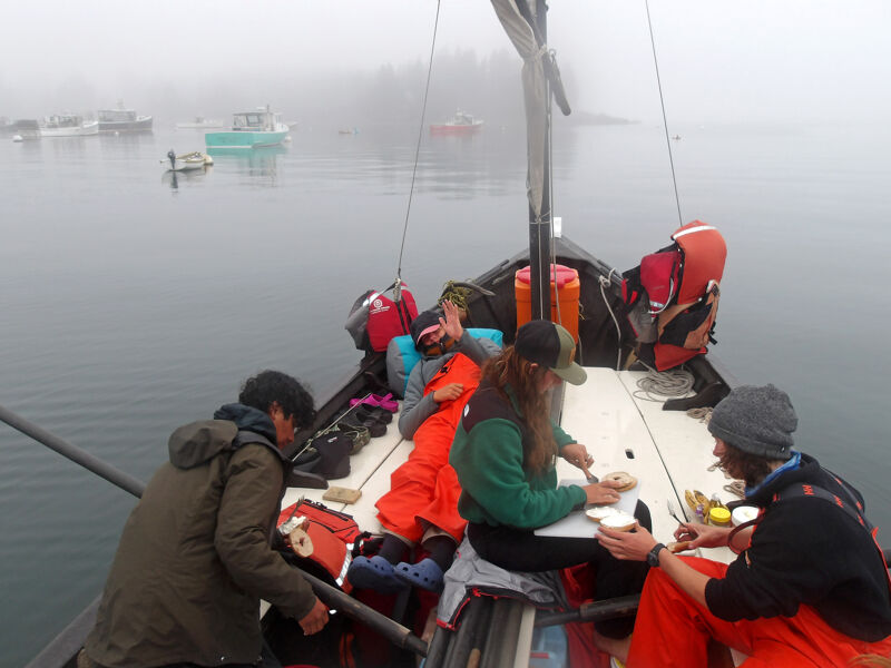 The image shows a group of people on a boat, possibly a small sailboat or fishing vessel, in foggy conditions. They appear to be eating or preparing food, with some wearing life jackets. The boat is surrounded by calm water, and other boats are visible in the distance through the fog. The overall scene suggests a peaceful, overcast day on the water.