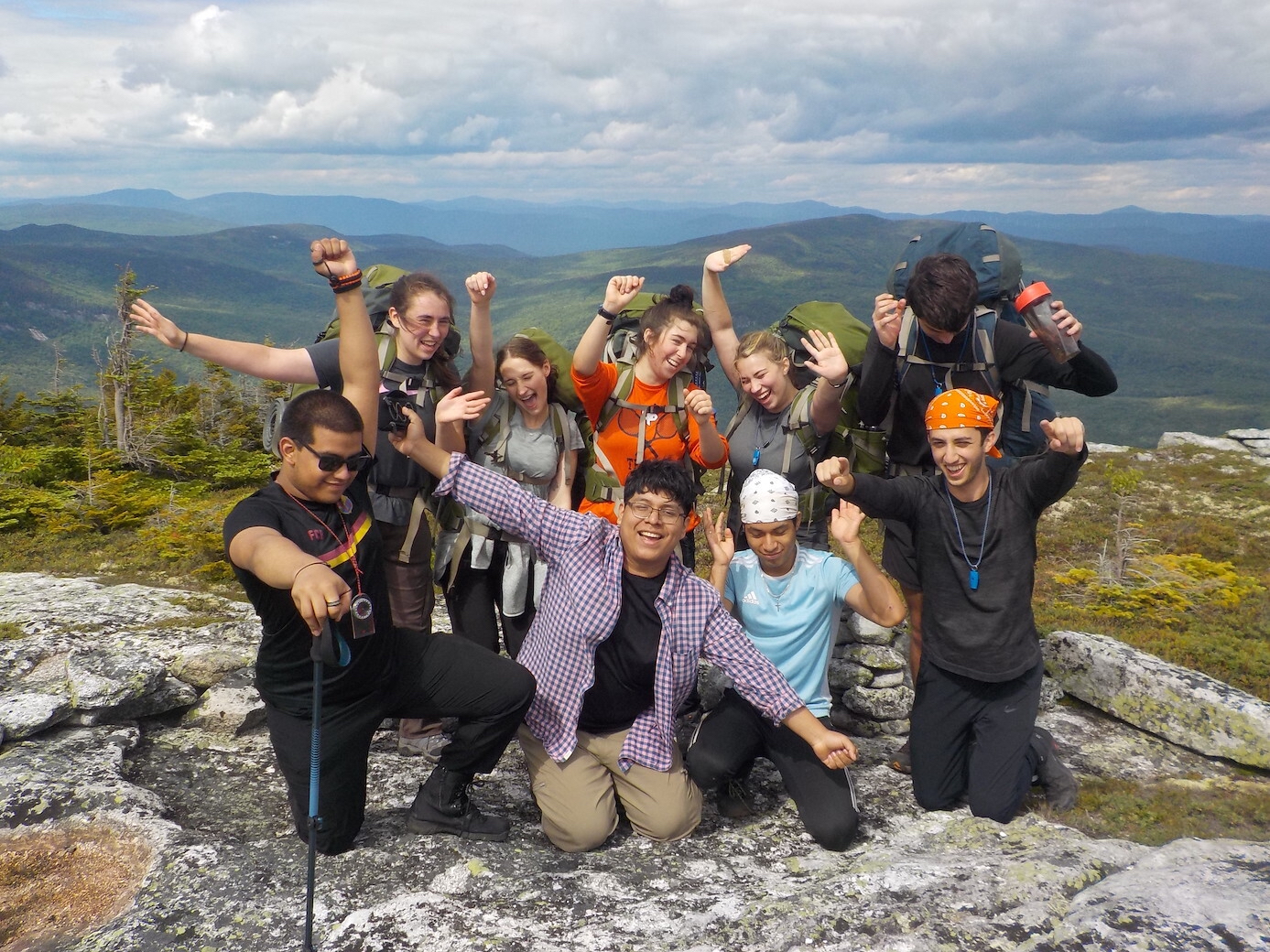 The image shows a group of young people on a mountaintop, celebrating with raised arms and joyful expressions. They are dressed in hiking gear, suggesting they have reached the summit after a hike. The background reveals a scenic view of mountains and a cloudy sky, enhancing the sense of accomplishment and adventure.