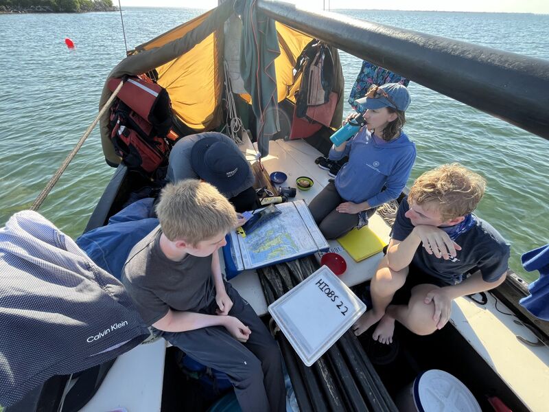 The image shows a group of people on a boat. They appear to be engaged in various activities, including looking at a map and drinking from a water bottle. The boat has a sail and some gear. The overall scene suggests a day of sailing or boating. The atmosphere seems relaxed and casual.
