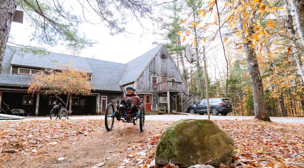 The image shows a person in a wheelchair in front of a wooden building. The building has a basketball hoop. There are trees with autumn leaves around the building. Another person in a wheelchair is visible in the background. A car is parked near the building.