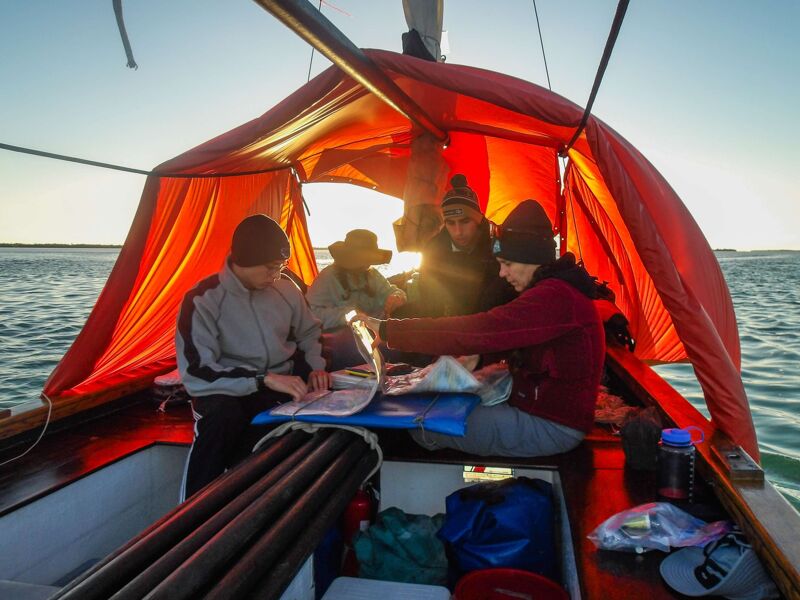 The image shows four people inside a small boat with a red canopy. They are looking at a map or chart, likely navigating. The sun is setting, casting a warm glow. The scene suggests a sailing adventure or a journey on the water.