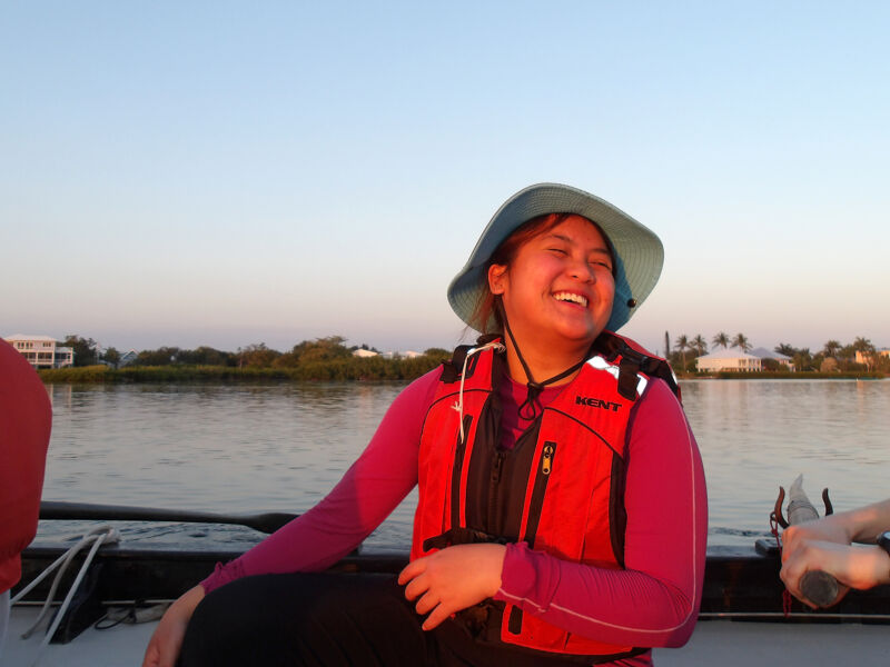 The image shows a young person on a boat, wearing a life jacket and a hat, and smiling broadly. The background includes water, trees, and a building, suggesting an outdoor setting, possibly during a sunset or sunrise. The overall mood is happy and relaxed.