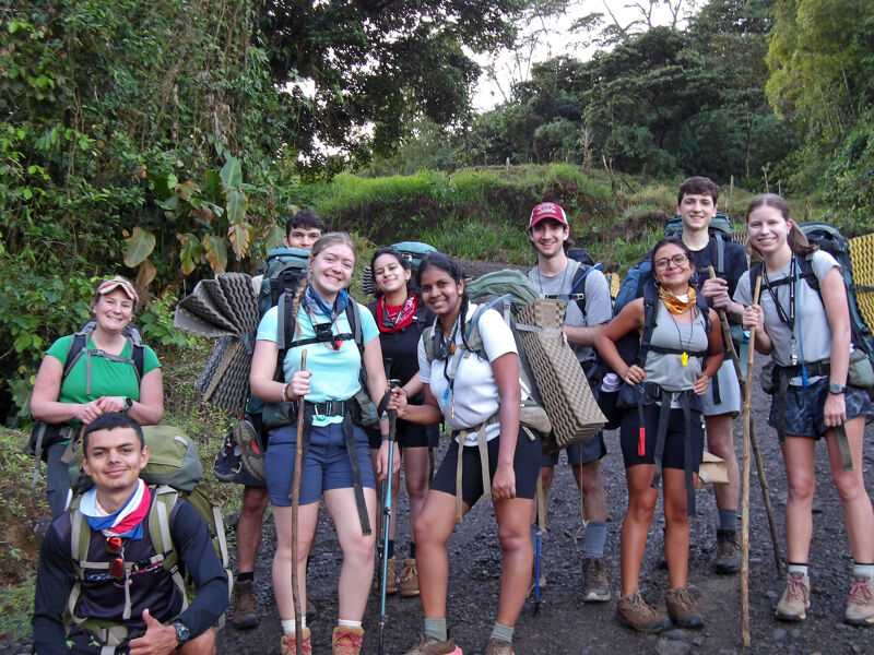 The image shows a group of young people, likely hikers, posing for a photo. They are in a natural setting, possibly a forest or a trail, with backpacks and hiking gear. The group seems diverse and ready for an adventure. The overall mood is positive and adventurous.