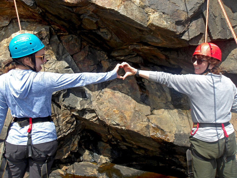 The image shows two people wearing helmets and climbing gear, likely rock climbing. They are extending their arms towards each other, forming a heart shape with their hands. They are smiling, suggesting they are enjoying the activity. The background is a rocky cliff face.