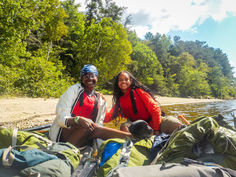 The image shows two smiling women sitting in a boat filled with backpacks. They are on a lake with trees in the background. The sky is blue with some clouds. They appear to be enjoying a day of outdoor adventure.