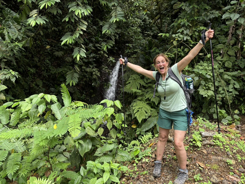 In the image, a woman is standing near a small waterfall in a lush, green forest. She is smiling and has her arms raised, appearing joyful. She is wearing hiking clothes and holding trekking poles, suggesting she is on a hike. The scene is vibrant and full of life.