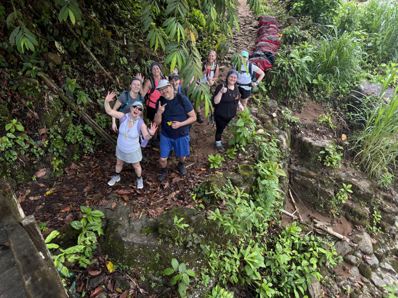 The image shows a group of people hiking on a trail in a lush, green environment. They are surrounded by dense vegetation, including trees and plants. The hikers appear to be enjoying their time, with some smiling and waving at the camera. The trail seems to be narrow and surrounded by nature.