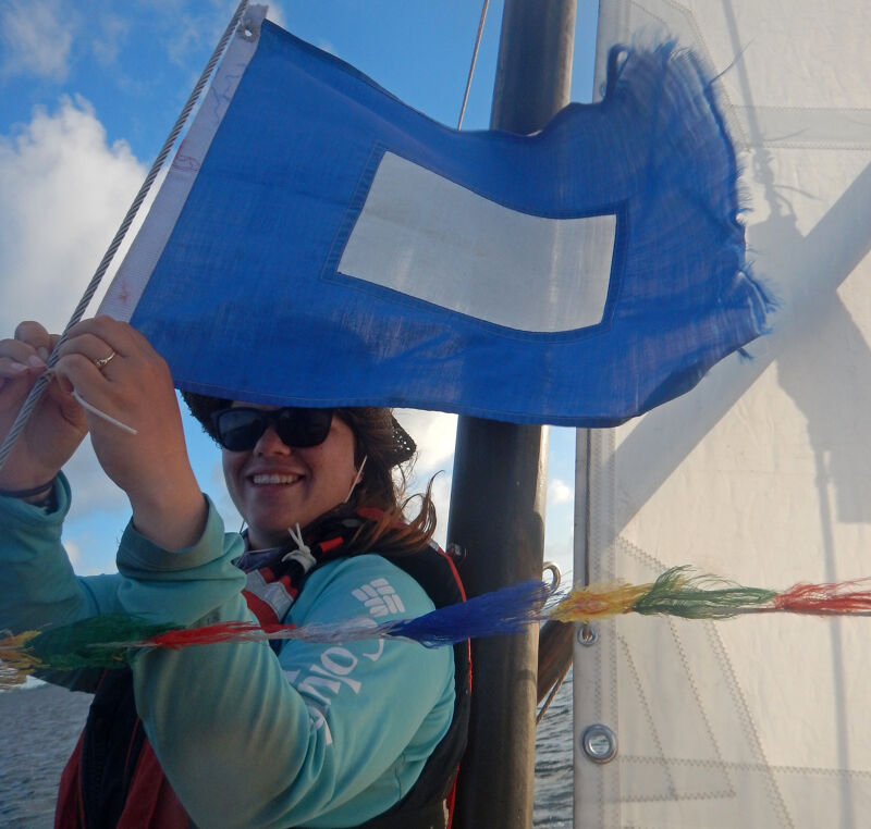 The image shows a person holding a blue flag with a white rectangle on it. The person is wearing sunglasses and a hat. The flag is being held up on a sailboat. The person appears to be smiling. The background shows a blue sky with some clouds.