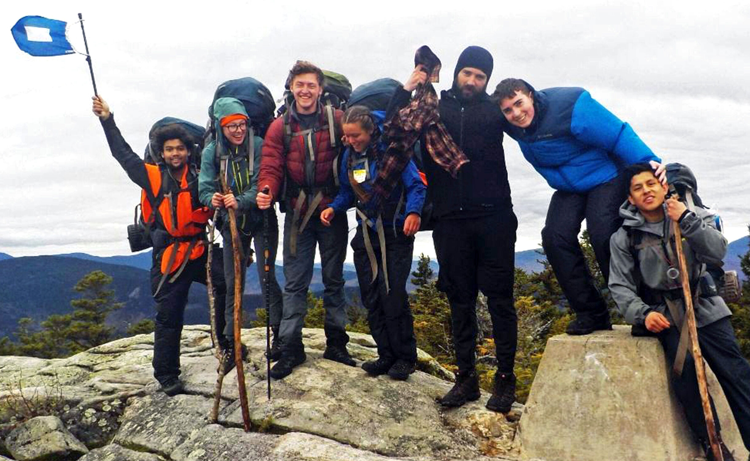 The image shows a group of hikers standing on a rocky mountaintop. They are all wearing backpacks and outdoor gear. One person is holding a flag. They appear to be celebrating their accomplishment. The sky is overcast, suggesting a cool day.