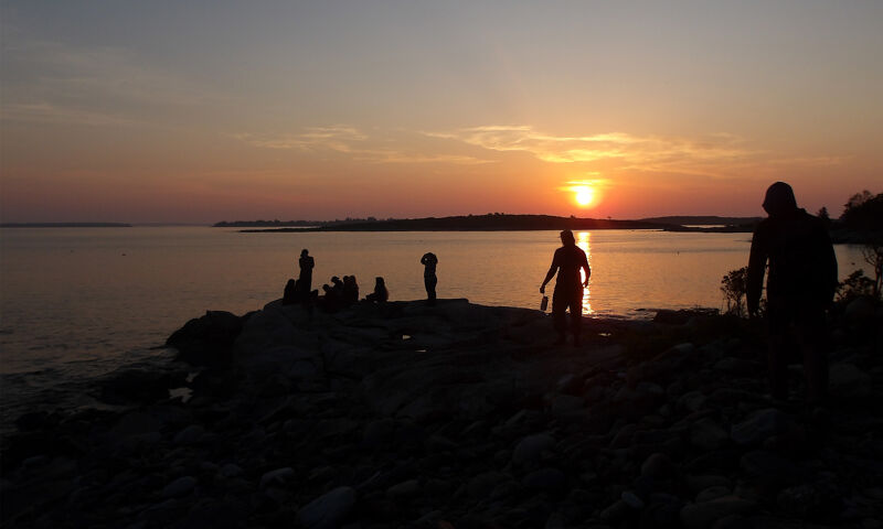 The image captures a serene sunset over a body of water. Silhouetted figures of people are scattered along the rocky shoreline, some seemingly watching the sunset. The sky is ablaze with warm hues of orange and yellow, reflecting on the water's surface. The overall atmosphere is peaceful and picturesque.