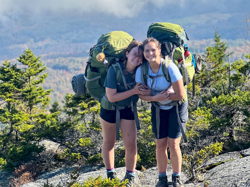 The image shows two young women smiling and embracing each other while standing on a rocky mountaintop. They are both wearing backpacks, suggesting they are on a hiking or camping trip. The background features a scenic view of mountains and trees under a cloudy sky. The overall impression is one of friendship and adventure.