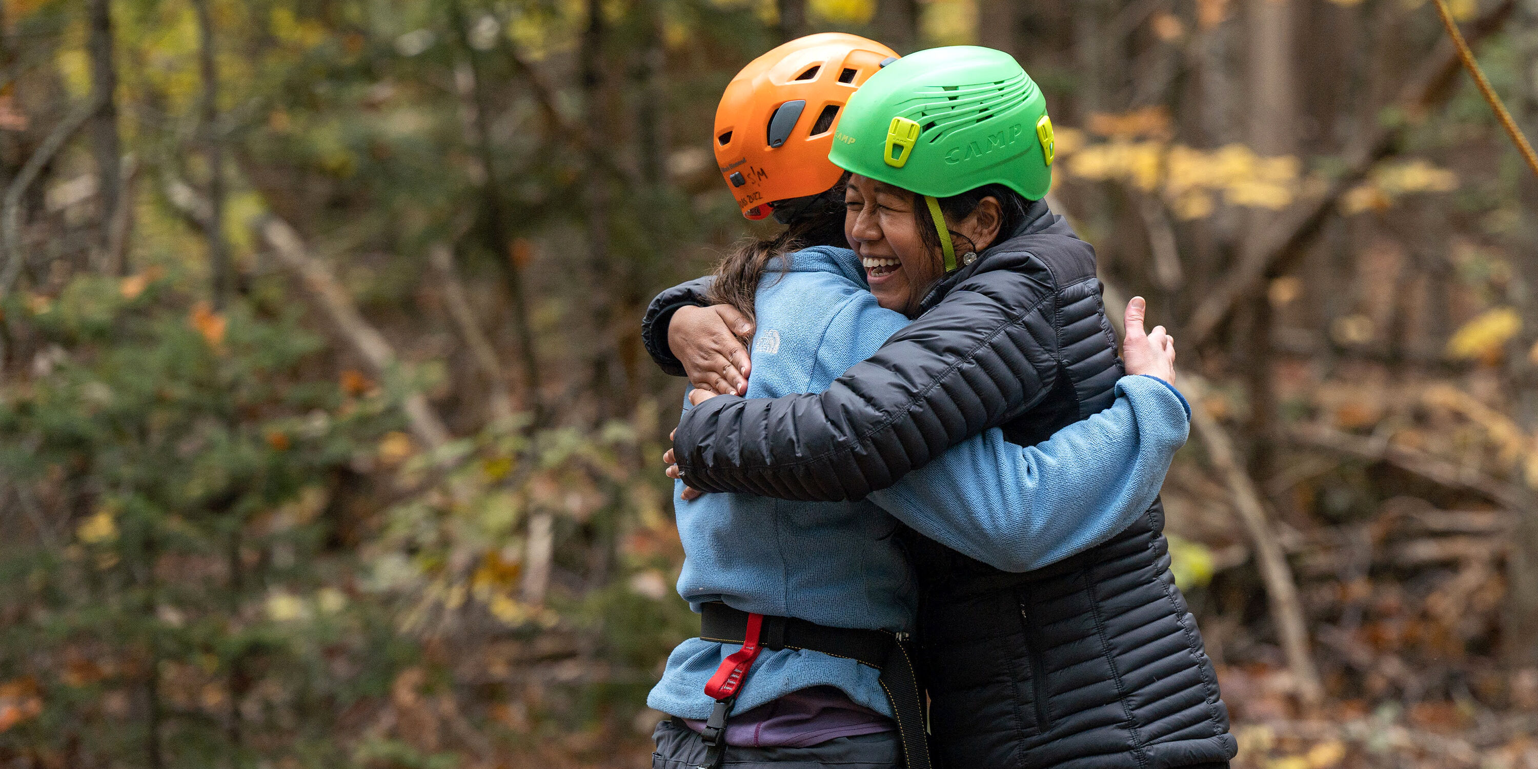 The image shows two people embracing each other. They are wearing helmets and outdoor gear, suggesting they are engaged in an activity like climbing or a ropes course. The background is a forest, with trees and foliage visible. The overall mood appears to be joyful and supportive.