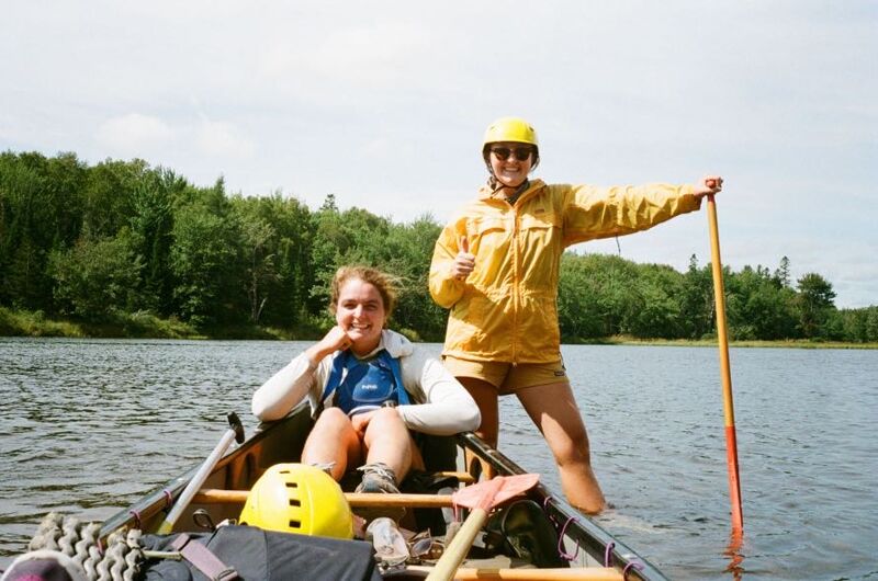 Here is a description of the image: Two young women are in a canoe on a lake. One is sitting and smiling, while the other is standing and holding a paddle. They are both wearing life jackets and helmets. The weather appears to be sunny.