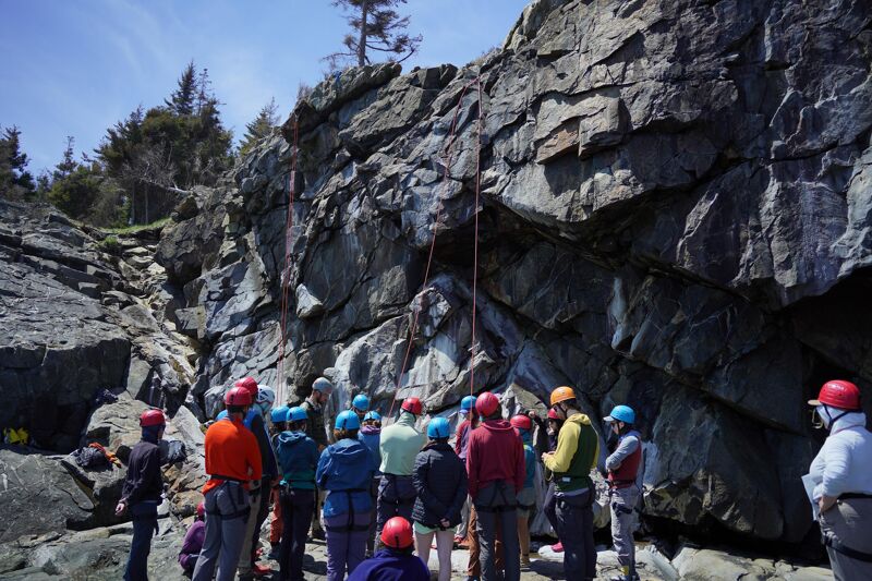 The image shows a group of people gathered at the base of a large rock formation. They are wearing helmets and appear to be preparing for or observing rock climbing. Ropes are visible, suggesting a climbing activity is underway. The setting is outdoors, with trees and a blue sky in the background.
