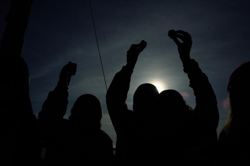 The image shows a group of people silhouetted against the sun. They are holding objects up in the air, possibly in celebration or offering. The bright sun creates a strong contrast with the dark figures, emphasizing their shapes and gestures. The overall impression is one of unity and shared experience.