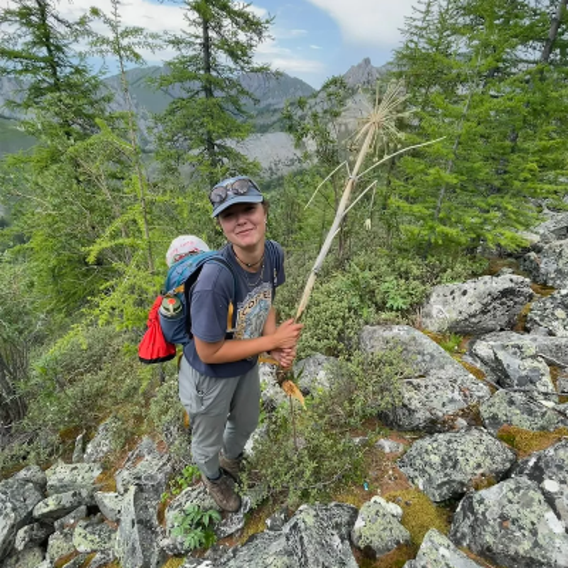 The image shows a woman and a baby in a carrier hiking in a mountainous area. The woman is holding a dried plant stalk. The background features trees, rocks, and a view of the mountains. The woman is smiling, and the overall scene suggests a day of outdoor adventure and exploration in nature.