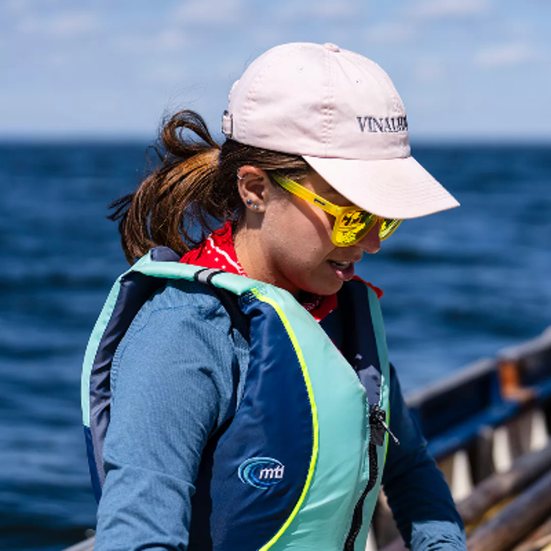 The image shows a woman wearing a pink cap, sunglasses, and a life jacket. She is looking down, possibly at something in the water. The background is the ocean, and the overall impression is of a sunny day on the water. She seems to be enjoying the outdoors.