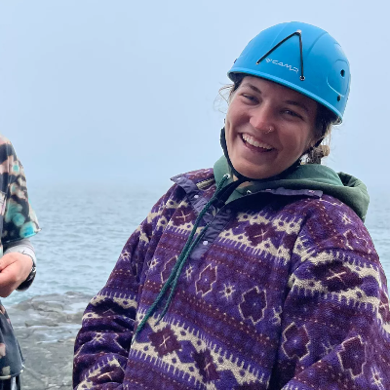 The image shows a person wearing a blue helmet and a purple patterned sweater. They are smiling and appear to be outdoors, possibly near a body of water, with a foggy background. The person seems happy and ready for an adventure.