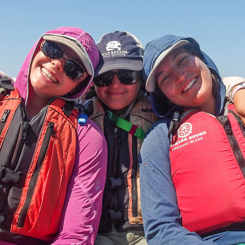 The image shows three smiling women wearing sunglasses, life jackets, and hooded jackets. They appear to be outdoors, possibly on a boat or near water, enjoying a sunny day. The woman in the middle is wearing a cap that says "Wild Rockies Field Institute". The woman on the right has a life jacket that says "Hurricane Island".