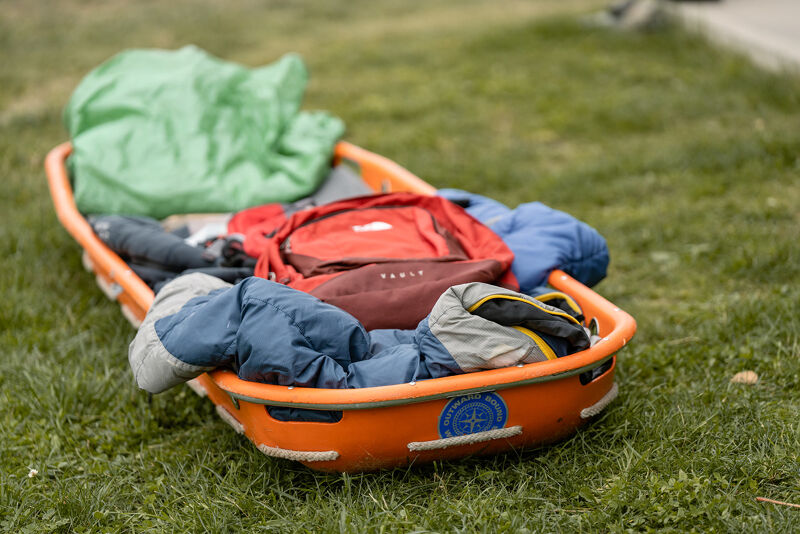 The image shows an orange sled filled with various items, including a green blanket, a red backpack, and several jackets. The sled is resting on a patch of green grass, suggesting an outdoor setting. The items appear to be packed for a trip or outdoor activity.
