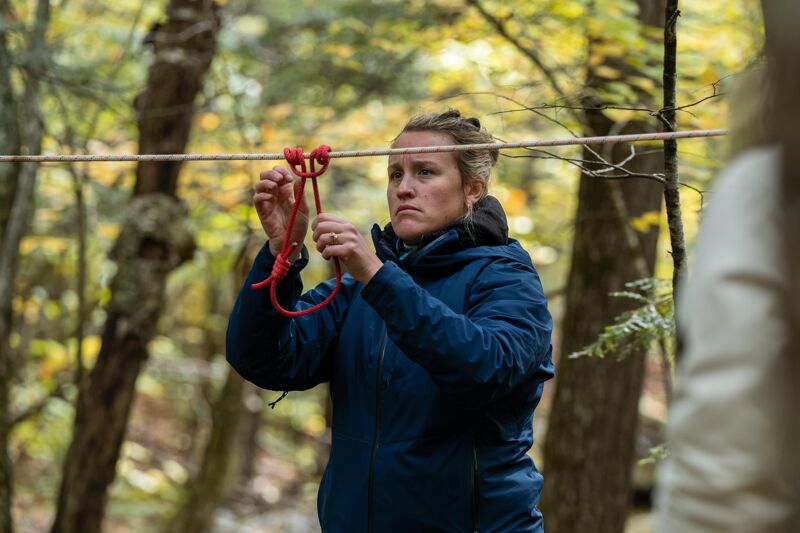 In the image, a woman is focused on manipulating a red rope attached to a horizontal line. She is wearing a blue jacket and appears to be outdoors, with a forest background. The scene suggests an activity involving rope work or a similar task. The woman's expression indicates concentration on the task at hand.