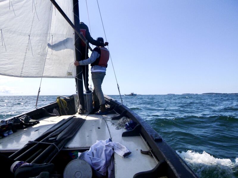 The image shows two people on a sailboat. One person is standing near the mast, and the other is further back. The boat is on the water, and the sail is up. The sky is blue, and the sea is choppy. In the distance, there is another boat and some land.
