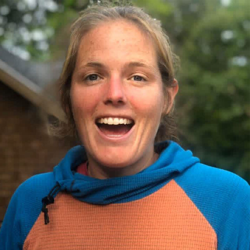 The image shows a close-up of a woman with blonde hair, wearing a blue and orange hooded sweatshirt. She has a wide, open-mouthed smile and appears to be outdoors, with greenery visible in the background. Her expression is joyful and energetic.