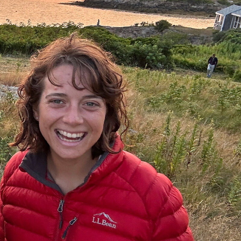 The image shows a close-up of a smiling woman with short, curly brown hair, wearing a red L.L.Bean puffer jacket. She is outdoors, with a grassy, hilly landscape and a body of water in the background. The lighting suggests it might be late afternoon or early evening. A person can be seen in the distance on the right side of the frame.