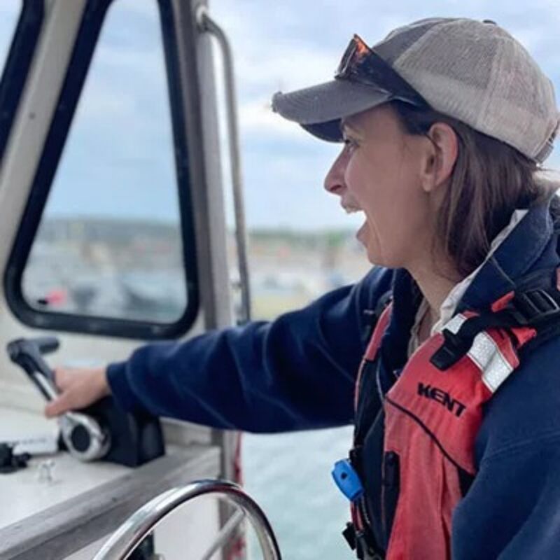 The image shows a woman on a boat, looking to the side with an excited expression. She is wearing a cap, a life jacket, and a dark blue jacket. Her hand is on the boat's controls. The background shows a body of water and a cloudy sky.
