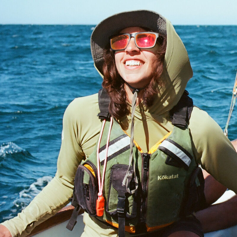 A woman with sunglasses and a hat is smiling while on a boat. She is wearing a life vest over a long-sleeved shirt. The water is blue and appears to be choppy. The sun is shining, and she seems to be enjoying herself on a sailing trip.