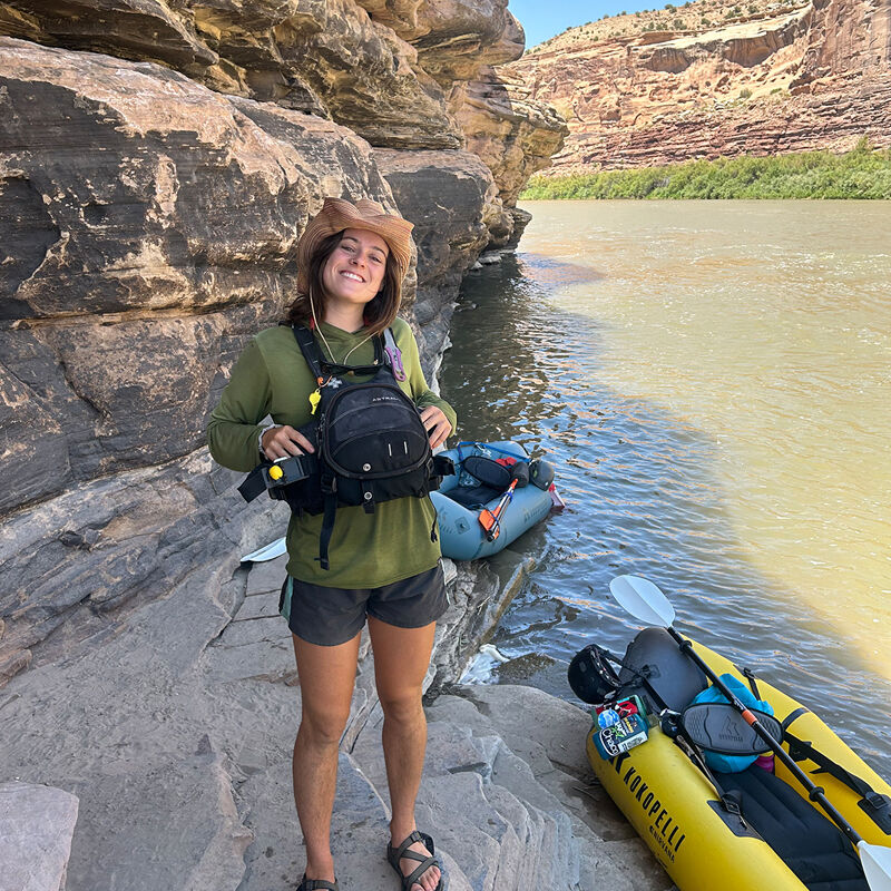 A young woman in a wide-brimmed hat, long-sleeved shirt, and shorts stands on a rocky shore next to a river. She is wearing a life vest and a backpack, and appears ready for an outdoor adventure. In the background, a raft and kayak are visible on the water, with a canyon wall rising behind them. The scene suggests a day of whitewater rafting or kayaking in a scenic, natural environment.