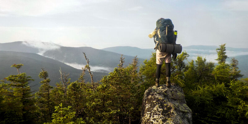 A hiker stands atop a rocky outcrop, gazing out at a vast, misty mountain range. The early morning sun casts a soft glow over the landscape, highlighting the layers of rolling hills and the dense evergreen forest. The hiker, equipped with a large backpack, appears to be taking a moment of quiet contemplation amidst the serene natural beauty.