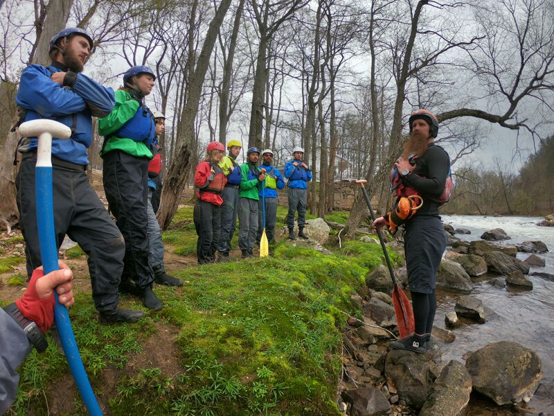 A group of people are gathered near a river, seemingly preparing for a water activity like kayaking or rafting. They are dressed in outdoor gear, including life jackets and waterproof clothing. Some are holding paddles, and they appear to be listening to a person who might be giving instructions or guidance before they head out onto the water. The setting is a wooded area with the river flowing alongside.