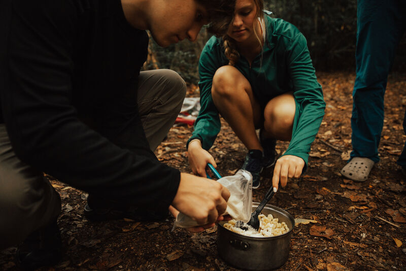 In a wooded area, two people are preparing food. One person, wearing a black long-sleeved shirt and khaki pants, is pouring liquid from a plastic bag into a pot. The other person, in a green jacket and dark shorts, is stirring the contents of the pot with a black utensil. The pot is sitting on the ground amidst fallen leaves and twigs.
