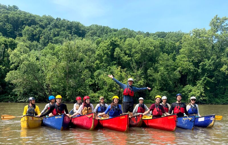 A group of people wearing life jackets and helmets are paddling in canoes on a river. They are surrounded by lush green trees and the sky is blue. One person in the middle canoe is raising their arms in the air, seemingly enjoying the activity. The canoes are brightly colored, adding a vibrant touch to the scene.