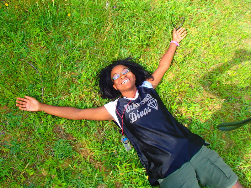 A young woman with dark hair and glasses is lying on her back in a grassy field. She is wearing a black jersey with the words "Diamond Divas" on it, along with khaki shorts. Her arms are outstretched, and she is smiling. The sun is shining, and the grass is green. The overall impression is one of joy and relaxation.