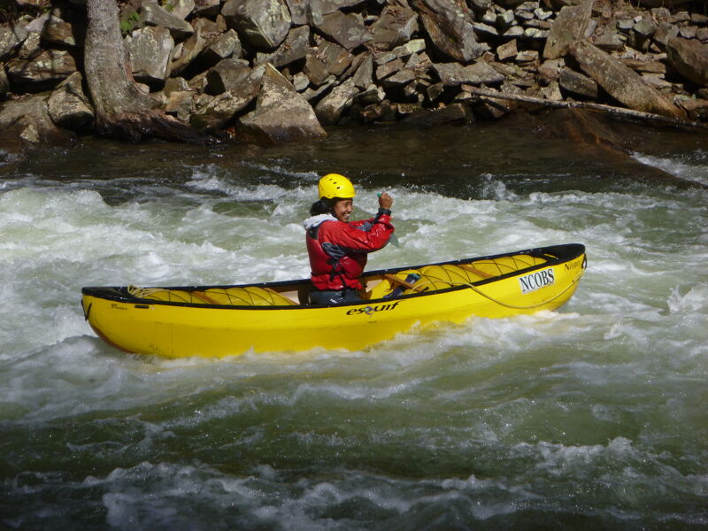 A person in a yellow kayak navigates through choppy water. The kayaker wears a yellow helmet and a red jacket. The kayak is surrounded by white water, indicating rapids or a fast-moving current. The background features a rocky shoreline with trees and foliage. The overall scene suggests an outdoor adventure activity, likely kayaking or canoeing in a natural river environment.