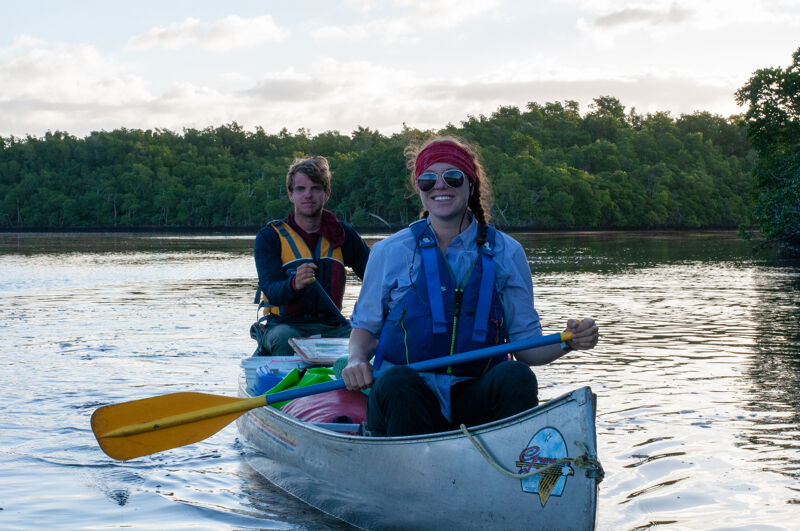 Two people are paddling a canoe on a body of water. The person in the front is wearing a red headband and sunglasses, and the person in the back is wearing a life jacket. The canoe is in the middle of the water, and there are trees in the background. The sky is cloudy.