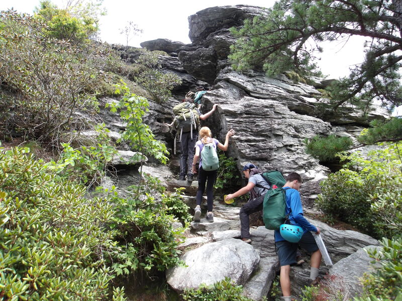 The image shows a group of people hiking up a rocky trail. They are surrounded by lush greenery and large rock formations. The hikers are wearing backpacks and appear to be enjoying their outdoor adventure. The sky is overcast, suggesting a cloudy day.
