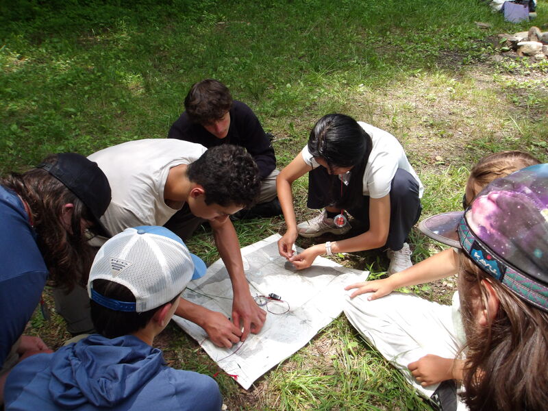 The image shows a group of young people gathered outdoors, likely engaged in a navigation or map-reading activity. They are clustered around a map spread on the ground, with some individuals pointing or examining it closely. The setting appears to be a grassy area, suggesting a recreational or educational context.