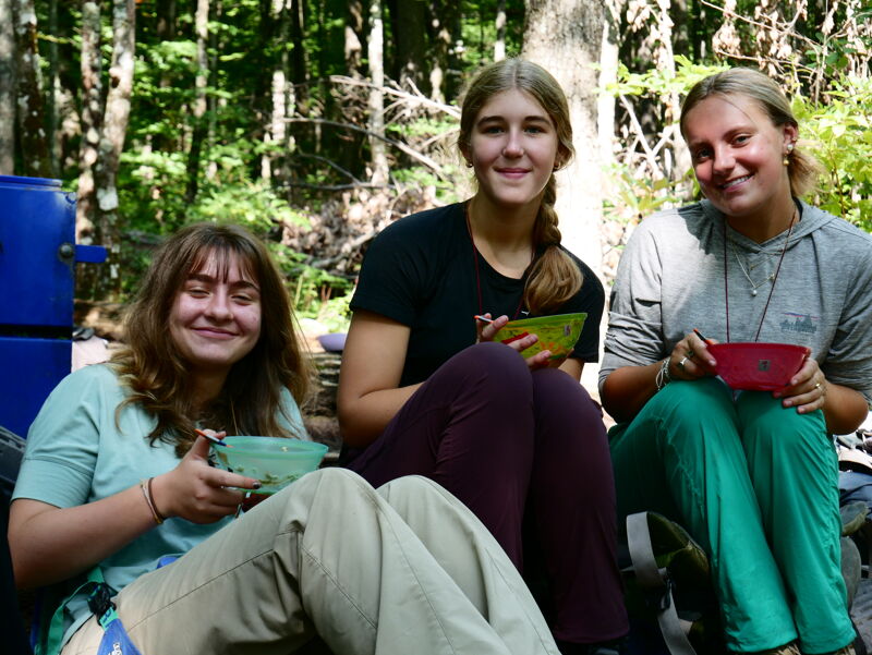 The image shows three young women sitting outdoors. They are smiling and appear to be enjoying themselves. They are holding bowls, possibly after a meal. The setting is a wooded area, suggesting they might be camping or on a nature outing.