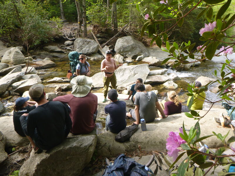 The image shows a group of people gathered near a stream in a natural setting. They appear to be on a hike or camping trip, with backpacks visible. Some individuals are sitting on rocks, while others are standing and interacting. The scene suggests a moment of rest or instruction amidst the outdoor activity.