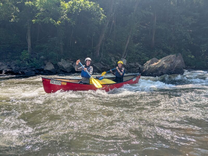 The image shows two people in a red canoe navigating a river with rapids. They are both wearing helmets and using paddles to steer the canoe. The water is turbulent, and the background features trees and rocks. The overall scene suggests an adventurous outdoor activity.