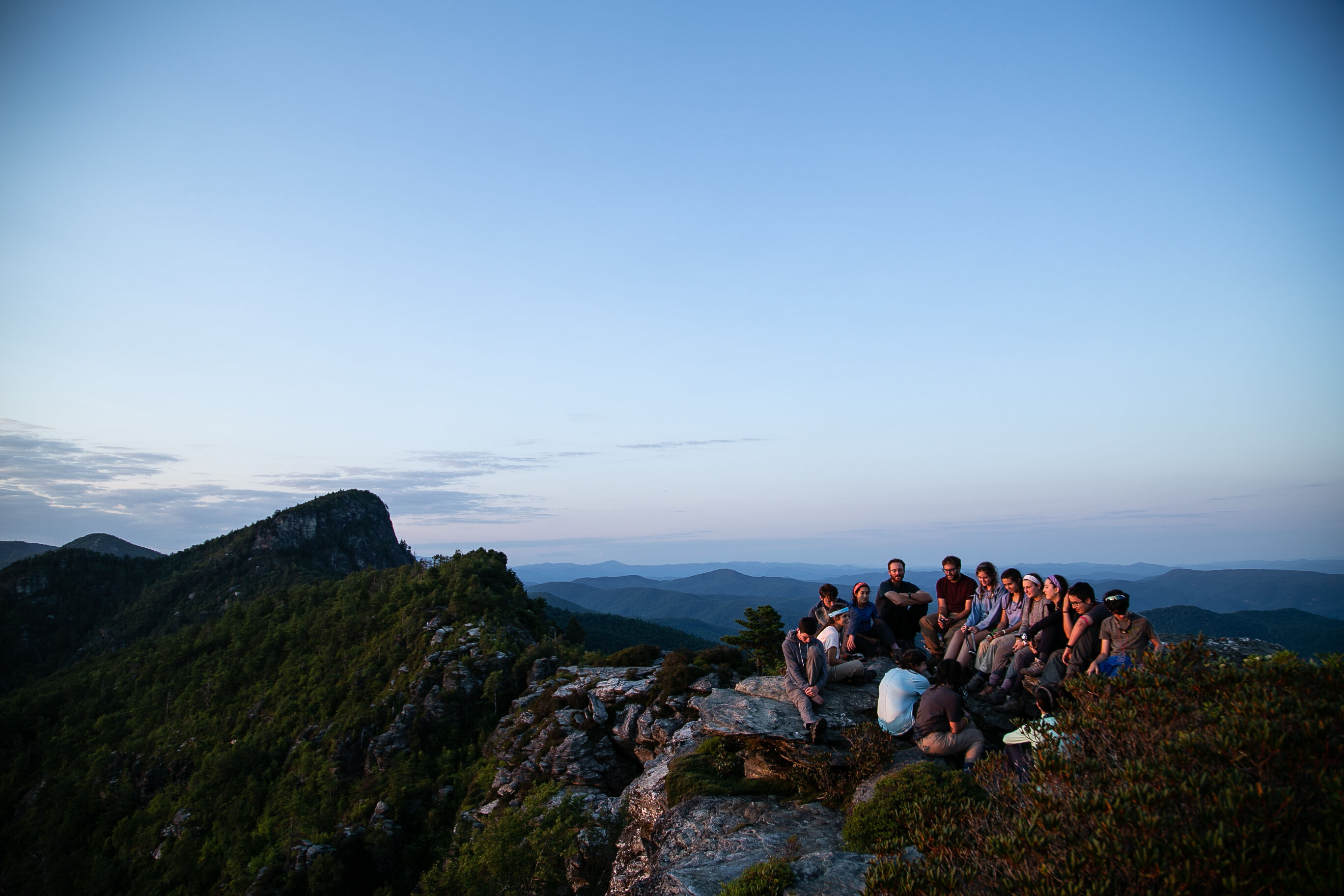 The image shows a group of people gathered on a rocky mountaintop at dusk or dawn. The sky is a gradient of blue, and the landscape features rolling hills and mountains covered in lush greenery. The group appears to be enjoying the scenic view and each other's company in a serene outdoor setting.