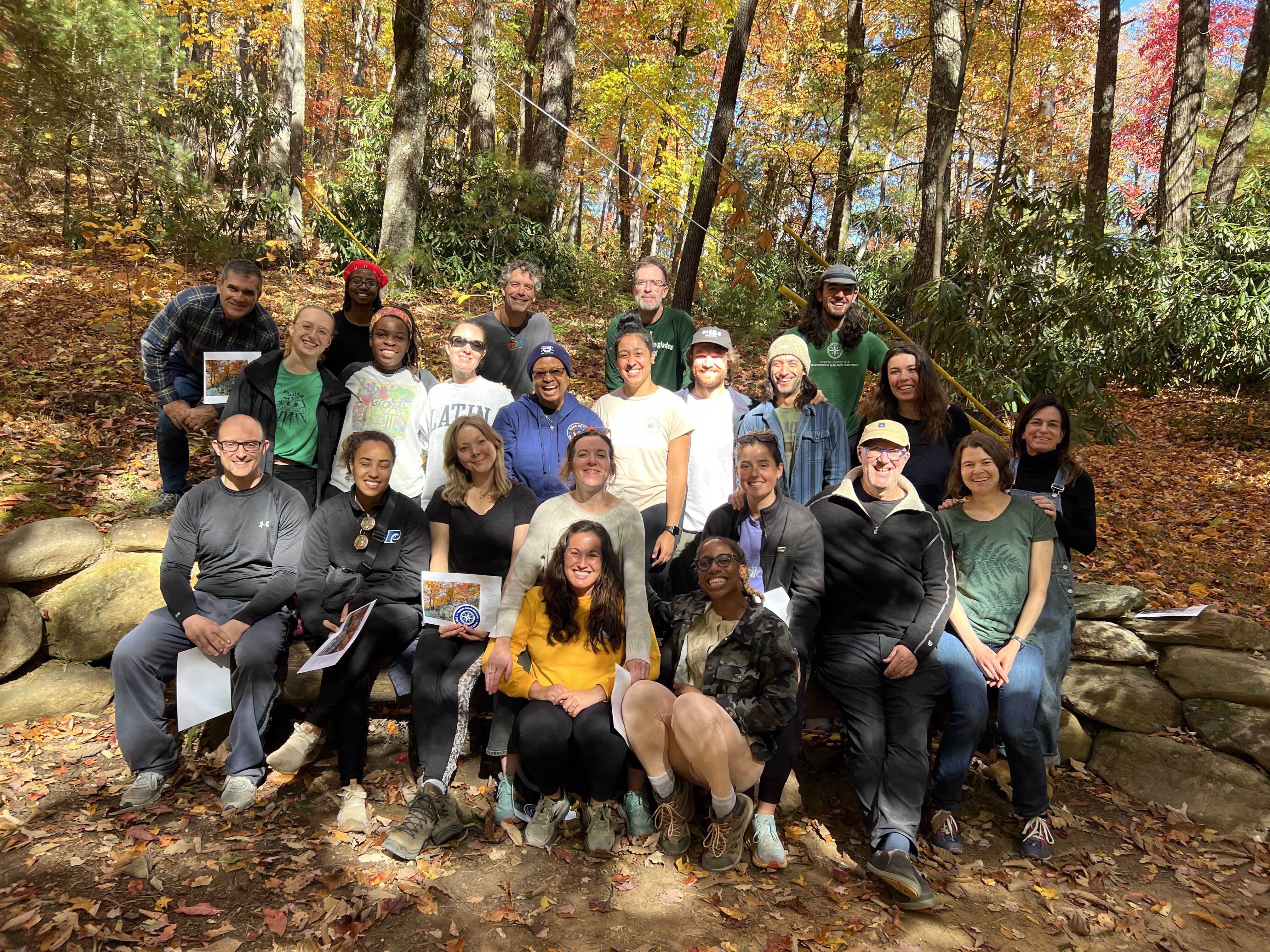 A diverse group of people, likely students and instructors, are gathered outdoors in a forest setting during autumn. They are posing for a group photo, with many smiling and looking at the camera. The trees display vibrant fall foliage in shades of yellow, orange, and red, creating a picturesque backdrop. Some individuals are holding papers or books, suggesting an educational or research activity.